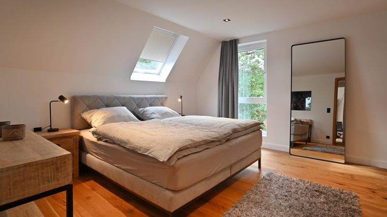 Ein modernes Schlafzimmer mit einem großen Bett und Holzfußboden. Es gibt ein Fenster mit Tageslicht und einen Spiegel an der Wand.