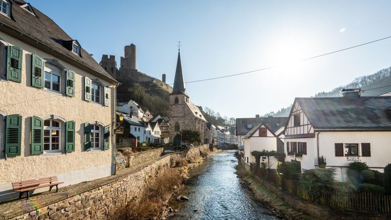 Eine malerische Stadt mit traditionellen Häusern an einem klaren Fluss. Im Hintergrund ist eine Kirche mit einem hohen Turm und eine Burg zu sehen.