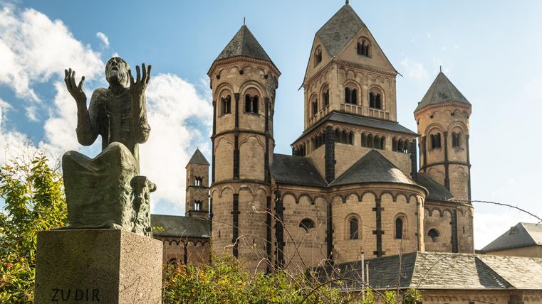 Eine historische Kirche mit charakteristischen Türmen steht im Hintergrund. Im Vordergrund befindet sich eine Statue, die eine betende Figur darstellt.