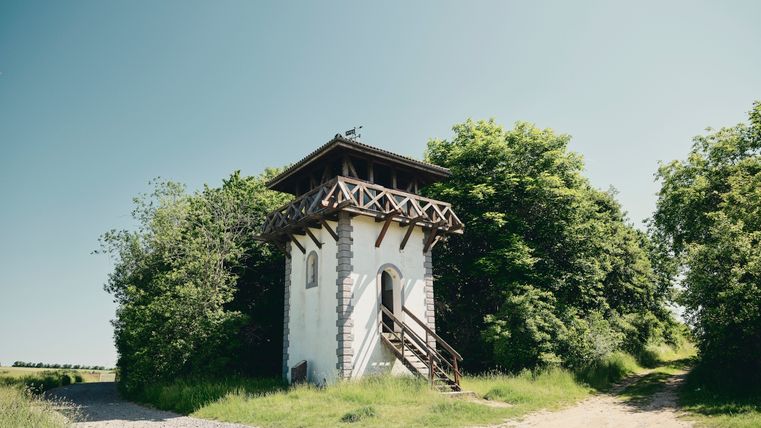 Ein rekonstruiertes römisches Wachhaus mit weißer Fassade, Holzbalkon und Treppe, umgeben von Bäumen und ländlichen Wegen unter klarem, blauem Himmel.