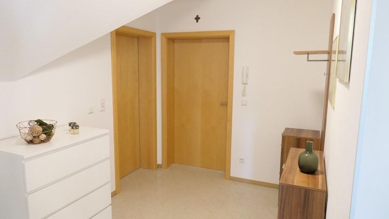 Een lichte gang met twee deuren en moderne meubels. Aan de linkerzijde staat een witte commode en aan de rechterzijde een houten commode met een vaas.