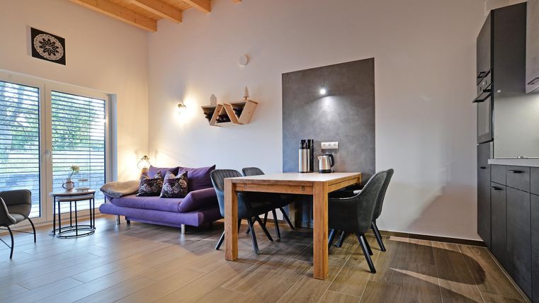 Ein modernes Wohn- und Esszimmer mit einem gemütlichen lila Sofa und einem Holz-Essstisch. Große Fenster sorgen für viel Tageslicht.