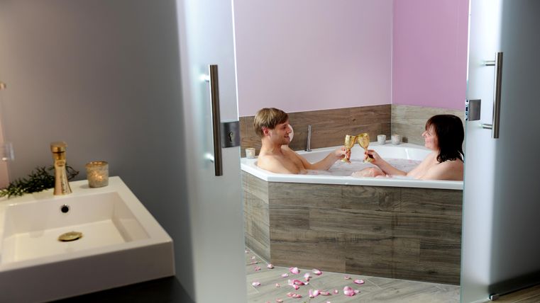 Ein Paar entspannt in einer Badewanne und stoßt mit Gläsern an. Um sie liegen Rosenblätter auf dem Boden, was eine romantische Atmosphäre schafft.