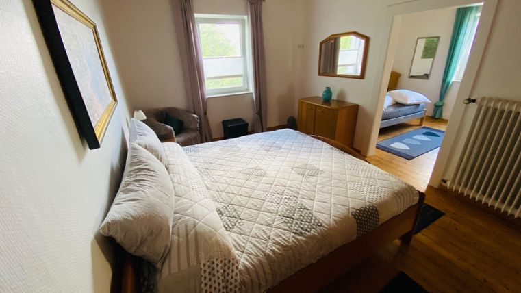 Ein gemütliches Schlafzimmer mit einem großen Bett und Holzfußboden. Die Wände sind hell, und es gibt Fenster, die viel natürlichem Licht hereinlassen.