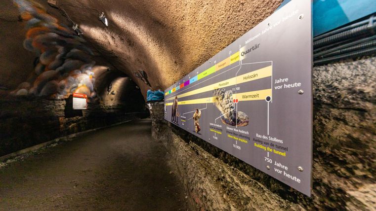 Ein unterirdischer Gang mit einer informationstafel an der Wand. Der Weg ist aus Stein und die Beleuchtung ist sanft.