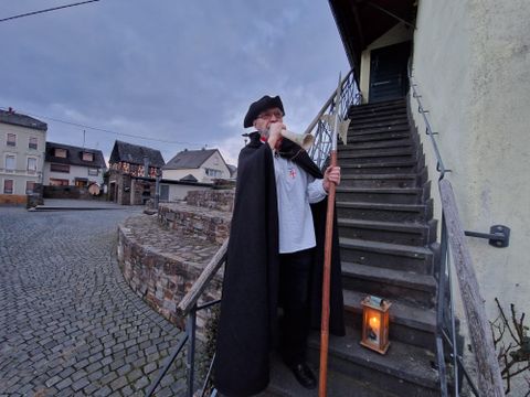 Ein Mann in einem schwarzen Umhang steht auf einer Treppe und hält einen Stock und eine Laterne. Im Hintergrund sind alte Gebäude und eine gepflasterte Straße zu sehen.