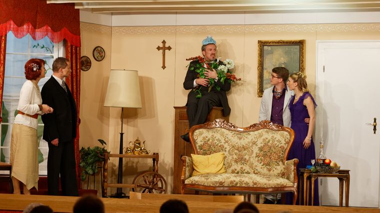Ein Schauspieler des Theatervereins Kaifenheim sitzt mit Blumen in den Händen auf einer Kommode, während die anderen vier Darsteller ihn während einer Szene umstehen.