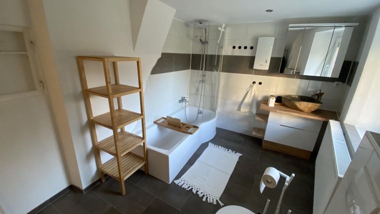 Ein modernes Badezimmer mit einer Badewanne und einer Dusche. Die Einrichtung ist hell und minimalistisch, mit einem Holzregal und einem sauberen, eleganten Design.