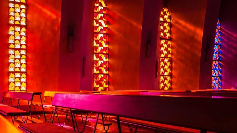 Ein schöner Kircheninnenraum mit farbigen Fenstern, die warmes Licht strahlen. Bänke aus Holz sind in der Mitte des Raumes aufgestellt.