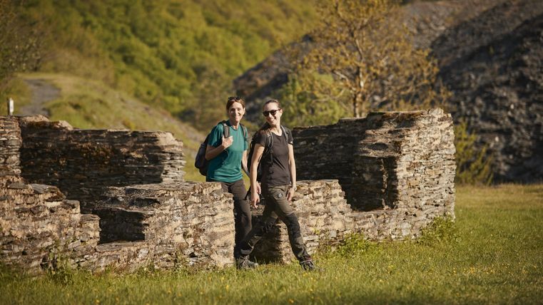 Zwei Personen stehen entspannt an einer historischen Steinmauer in einer grünen Landschaft. Die Umgebung ist von sanften Hügeln und Bäumen umgeben.