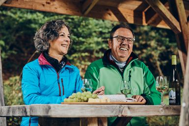 Zwei Menschen sitzen draußen an einem Holztisch, trinken Weißwein und essen Trauben in einer naturnahen Umgebung.