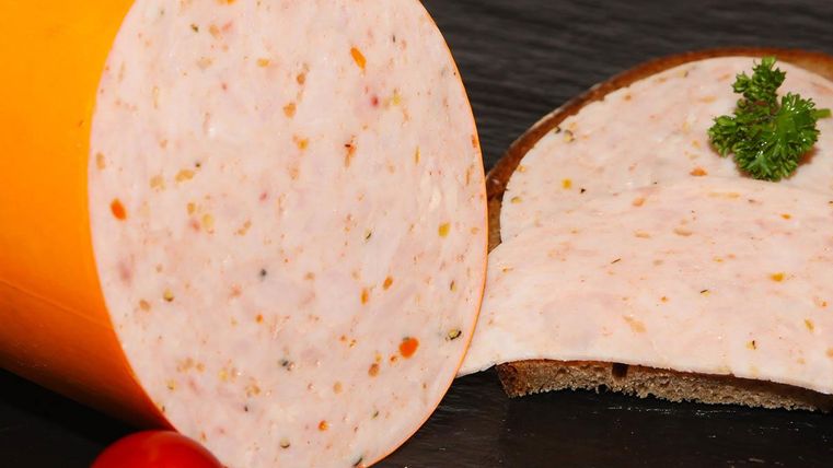 Eine runde Wurst mit bunten Einlagen liegt auf einem schwarzen Untergrund. Daneben sind einige Scheiben Wurst auf Brot und eine kleine Tomate zu sehen.