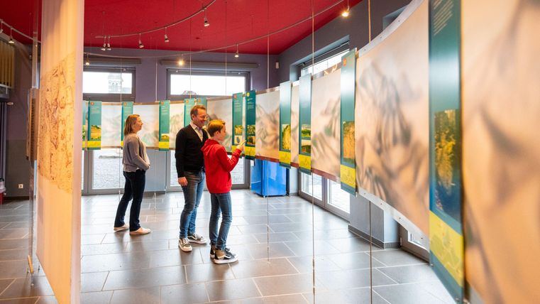 Eine Ausstellung in einem Raum mit mehreren Informationsplakaten. Besucher schauen sich die Kunstwerke an und interagieren mit den Exponaten.