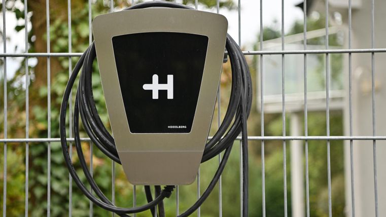 Eine Ladestation für Elektrofahrzeuge mit einem Kabel, das bereit zum Aufladen ist. Sie befindet sich hinter einem Zaun, umgeben von grüner Vegetation.