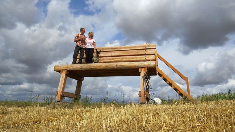 Große XXL-Holzbank mit Blick auf die Eifellandschaft bei Möntenich.Auf der linken Seite der Bank hat ein Mann eine Frau im Arm und daneben steht ein kleiner Hund.