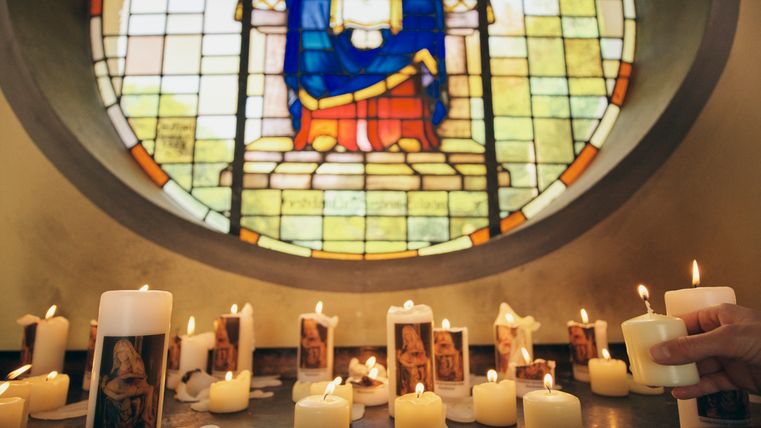 Brennende Kerzen mit Marienabbildungen vor einem farbenprächtigen Kirchenfenster mit religiösem Motiv; eine Hand stellt eine weitere Kerze dazu.