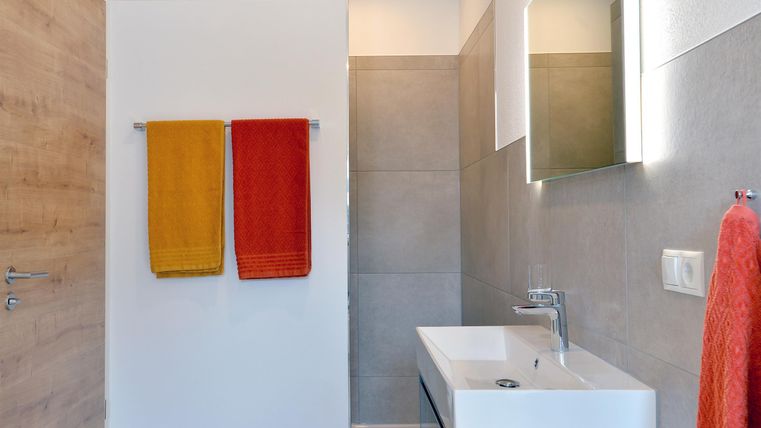 Ein modernes Badezimmer mit einer eleganten Waschbeckenanlage. Rote und orange Handtücher hängen an der Wand, während die Wände hell und neutral gestaltet sind.