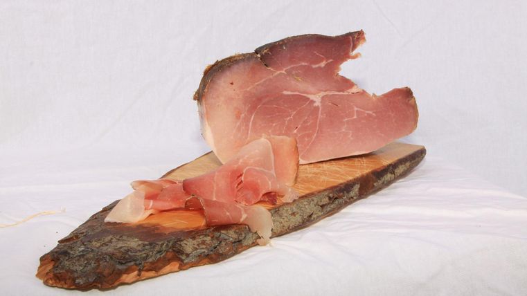 Ein Stück Schinken auf einem Holzbrett. Daneben liegen weitere geschnittene Schinkenscheiben.