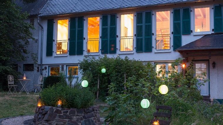 Une maison lumineuse et confortable avec des volets verts et des fenêtres éclairées. Au premier plan, il y a un jardin avec des plantes et des lumières ressemblant à des lanternes.