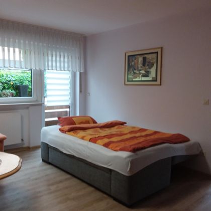 Ein gemütliches Schlafzimmer mit einem großen Bett und bunten Bettlaken. Das Zimmer hat ein Fenster mit Blick ins Grüne und einen kleinen Tisch.