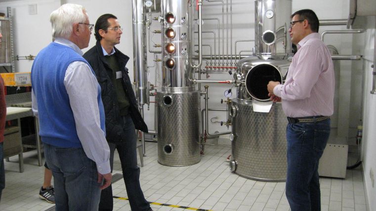 Een groep mannen luistert naar een lezing over een distillatie-installatie. Op de achtergrond zijn grote, glanzende roestvrijstalen apparaten te zien.