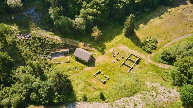 Eine kleine Holzstruktur liegt inmitten von üppigem grünen Wald. Umgeben von den Überresten anderer Gebäude und einer offenen Fläche.