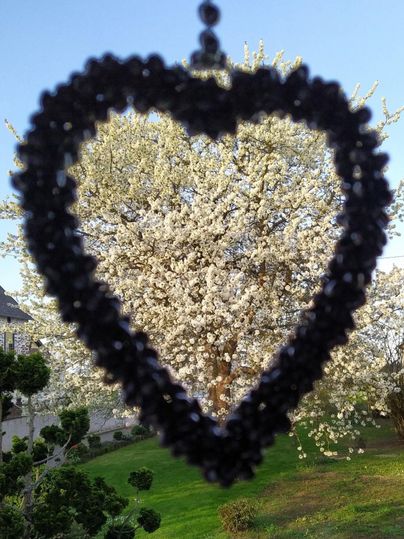 Ein herzförmiger Anhänger im Vordergrund, mit einem blühenden Baum im Hintergrund. Die Szenerie strahlt eine romantische Frühlingsatmosphäre aus.