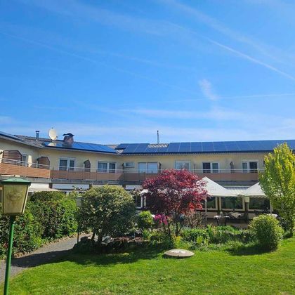Ein hübsches Hotel mit einem schönen Garten und bunten Pflanzen. Der Himmel ist klar und blauer Sonnenschein strahlt auf das Gebäude.