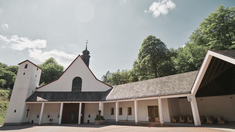 Eine moderne Kirche mit einem schlichten weißen Äußeren und einem gedeckten Dach. Umgeben von Bäumen unter einem blauen Himmel.