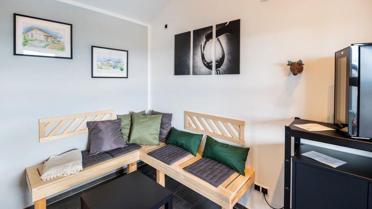 Ein gemütlicher Wartebereich mit Holzbänken und Kissen. An der Wand hängen Bilder, und ein kleines Möbelstück steht neben einem Kühlschrank.