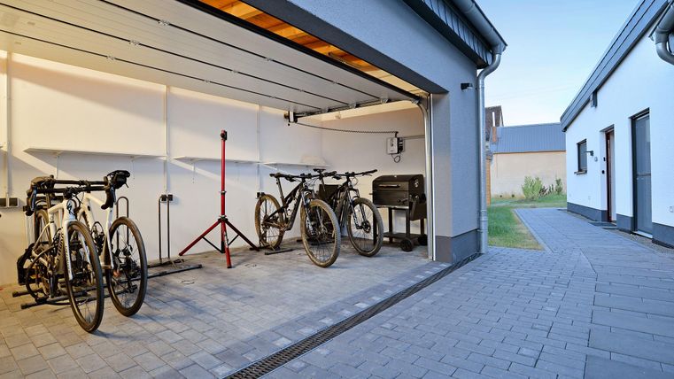 Eine moderne Garage mit mehreren Fahrrädern und einem Grill. Der Boden ist gepflastert und es gibt Zugang zu einem Nebengebäude.