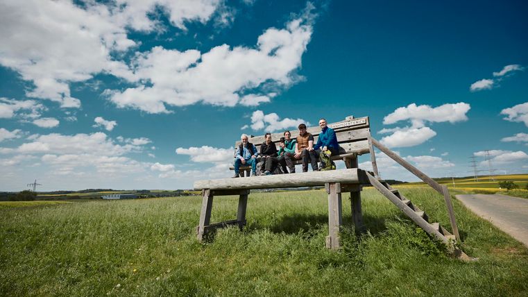 Eine Gruppe von Menschen sitzt auf einer Bank auf einem Hügel. Der Himmel ist blau mit ein paar Wolken, und die Landschaft ist grün.