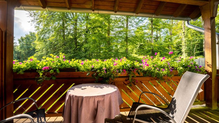 Ein gemütlicher Balkon mit einem runden Tisch und einem Stuhl. Bunte Blumen schmücken das Geländer und umgeben die grüne Landschaft.