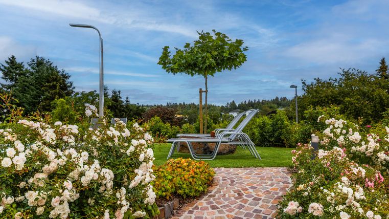 Ein schöner Garten mit Rosenblüten und einer gepflasterten Gehweg. Zwei Liegen stehen unter einem Baum, umgeben von üppigem Grün.