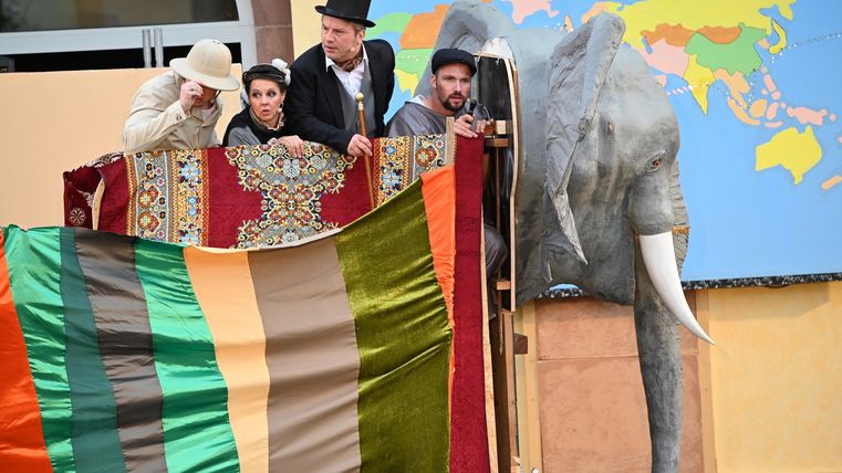 Darsteller des Theaterverein Kaisersesch spielen eine Szene aus „In 80 Tagen um die Welt“: Vier Figuren in historischen Kostümen blicken gespannt über einen bunt behängten Korb, der einen Heißluftballon darstellt, während neben ihnen ein großer Elefantenkopf als Bühnenrequisite und eine Weltkarte im Hintergrund zu sehen sind.