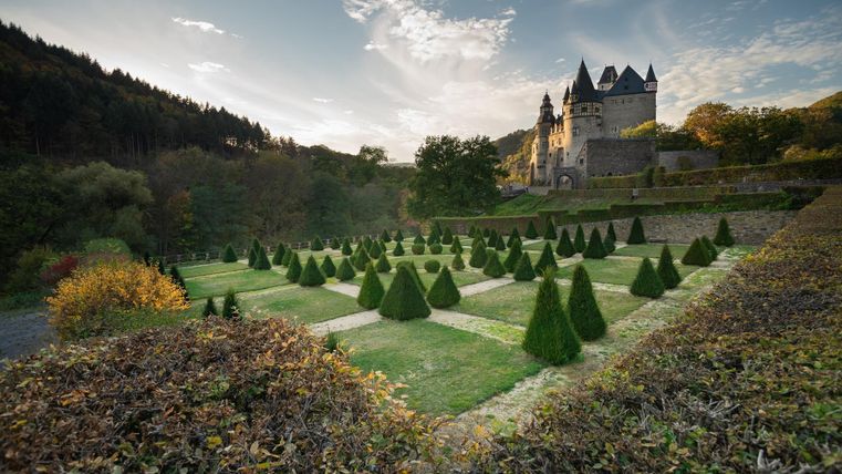 Eine malerische Landschaft mit einem Schloss im Hintergrund. Im Vordergrund sind geometrisch angeordnete, gepflegte Hecken zu sehen.
