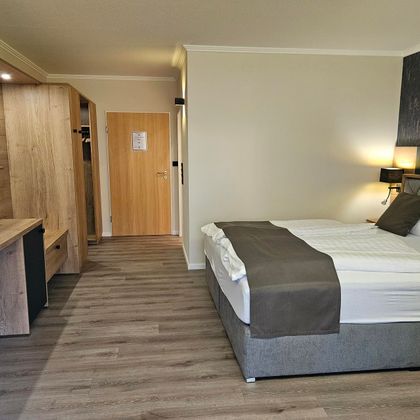 Ein modernes Hotelzimmer mit einem großen Bett und einem Schreibtisch aus Holz. Die Wände sind hell gestrichen, und der Boden ist mit Laminat ausgelegt.