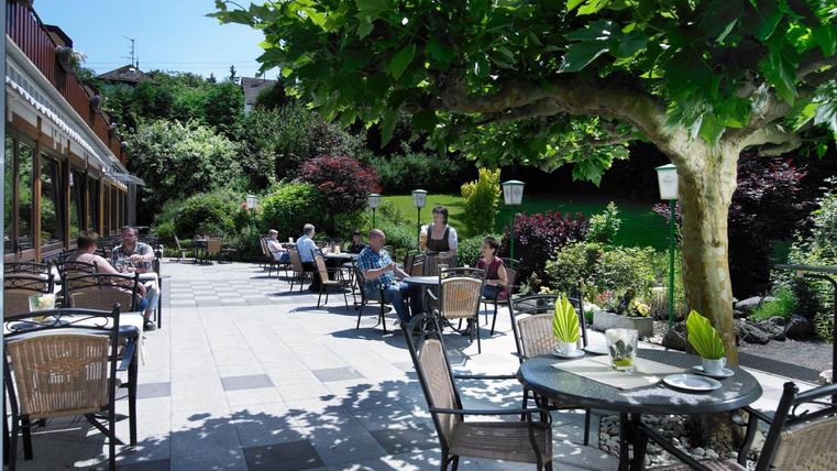 Ein einladender Außenbereich mit Tischen und Stühlen unter einem großen Baum. Gärten umgeben die Terrasse, und Gäste genießen die sonnige Atmosphäre.
