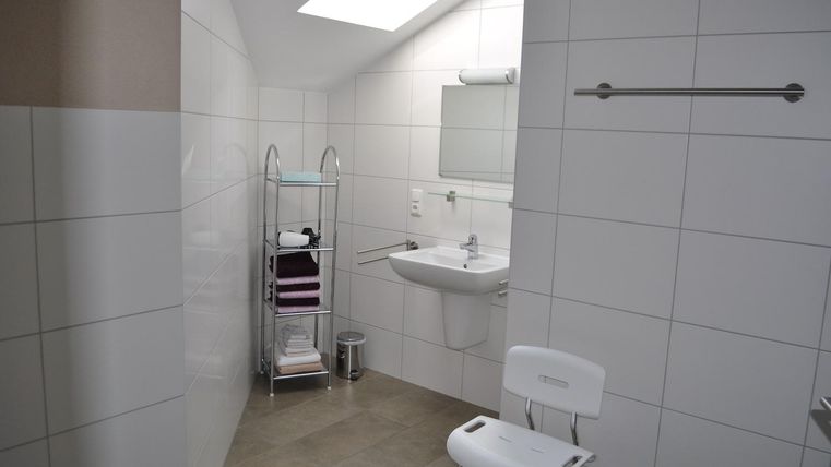 Ein modernes Badezimmer mit weißen Fliesen und einem großen Spiegel. Es gibt eine Aufbewahrungseinheit für Handtücher und eine Sitzgelegenheit.