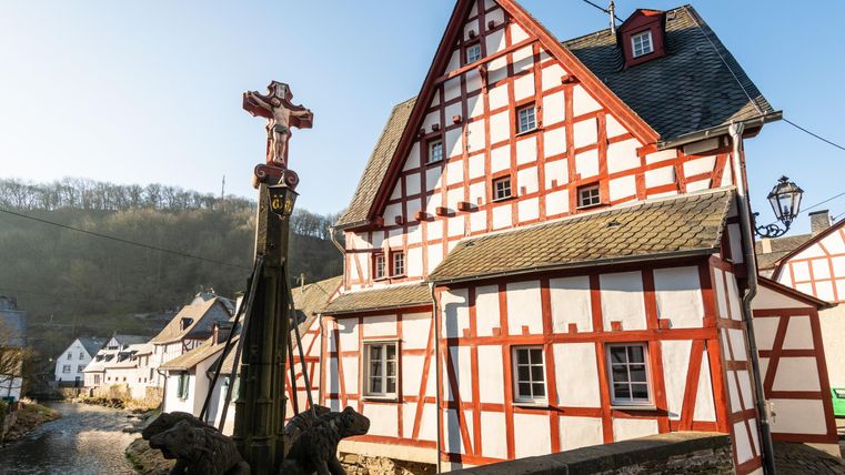 Ein historisches Fachwerkhaus mit roten und weißen Wänden steht an einem ruhigen Fluss. Im Vordergrund befindet sich ein Kreuz und eine Brücke, die den Blick auf die malerische Umgebung lenkt.