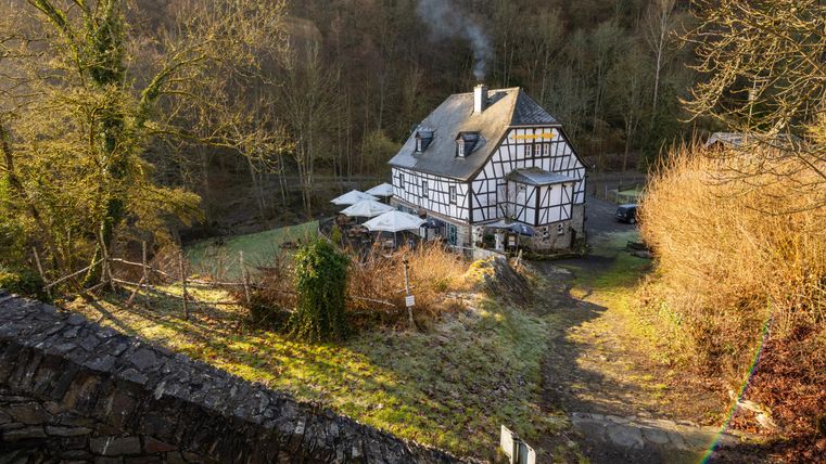 Ein malerisches Fachwerkhaus umgeben von Bäumen und sanften Hügeln. Rauch steigt aus dem Schornstein empor und das Wetter wirkt einladend und ruhig.