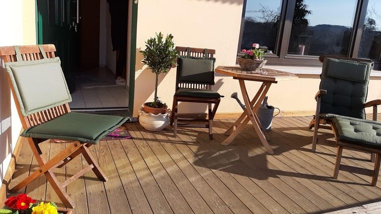 Een overdekt terras met een houten vloer en verschillende stoelen. Op de tafel staan bloemen en potten met kleurrijke planten.