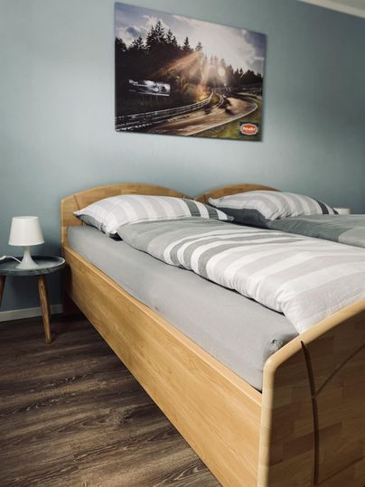Ein modernes Schlafzimmer mit einem hellen Holzbett und gestreiften Bettdecken. An der Wand hängt ein Bild einer Landschaft mit Sonnenstrahlen.