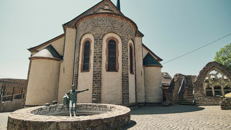 Eine historische Kirche mit einem runden Turm und einem schönen Mauerwerk. Im Vordergrund steht ein Brunnen mit einer Statue.