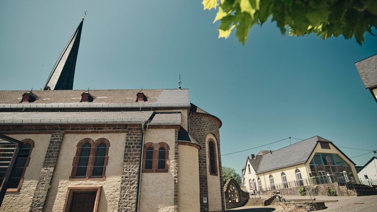 Eine alte Kirche mit einem hohen Turm und charakteristischen Fenstern. Im Hintergrund sieht man einige Häuser unter einem klaren blauen Himmel.