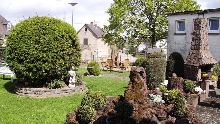 Een mooie tuin met een ronde struik, een stenen beeld en een zithoek. Op de achtergrond staat een barbecueplaats en er zijn veel decoratieve planten.