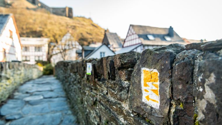 Wanderweg-Markierung auf einer Steinmauer mit Fachwerkhäusern im Hintergrund.