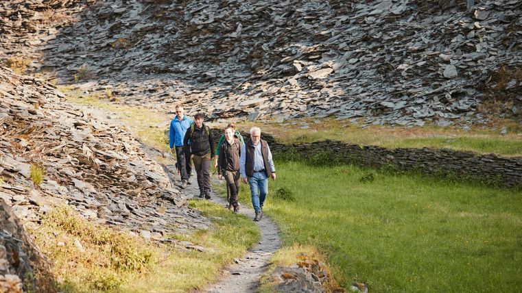 Een groep van vijf personen wandelt langs een smal pad, omgeven door stenen hellingen. Het landschap is groen en nodigt uit tot een verkenningstocht.