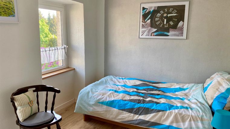 Ein gemütliches Schlafzimmer mit einem Bett und einer blauen Decke. Ein Fenster lässt viel Tageslicht herein und bietet einen Blick ins Grüne.