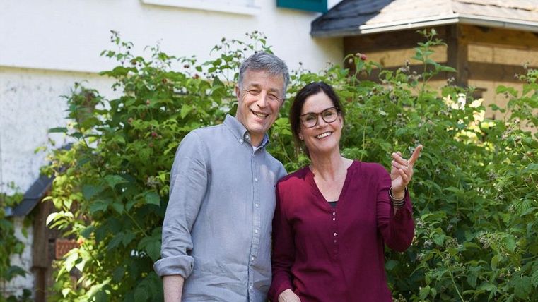 Un couple heureux se tient en souriant devant une maison, entouré de plantes vertes. Ils ont l'air détendu et amical.